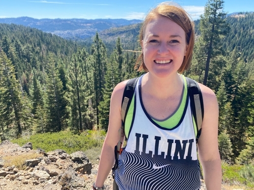 Photo of a woman smiling with a tree-filled mountain vista behind her.