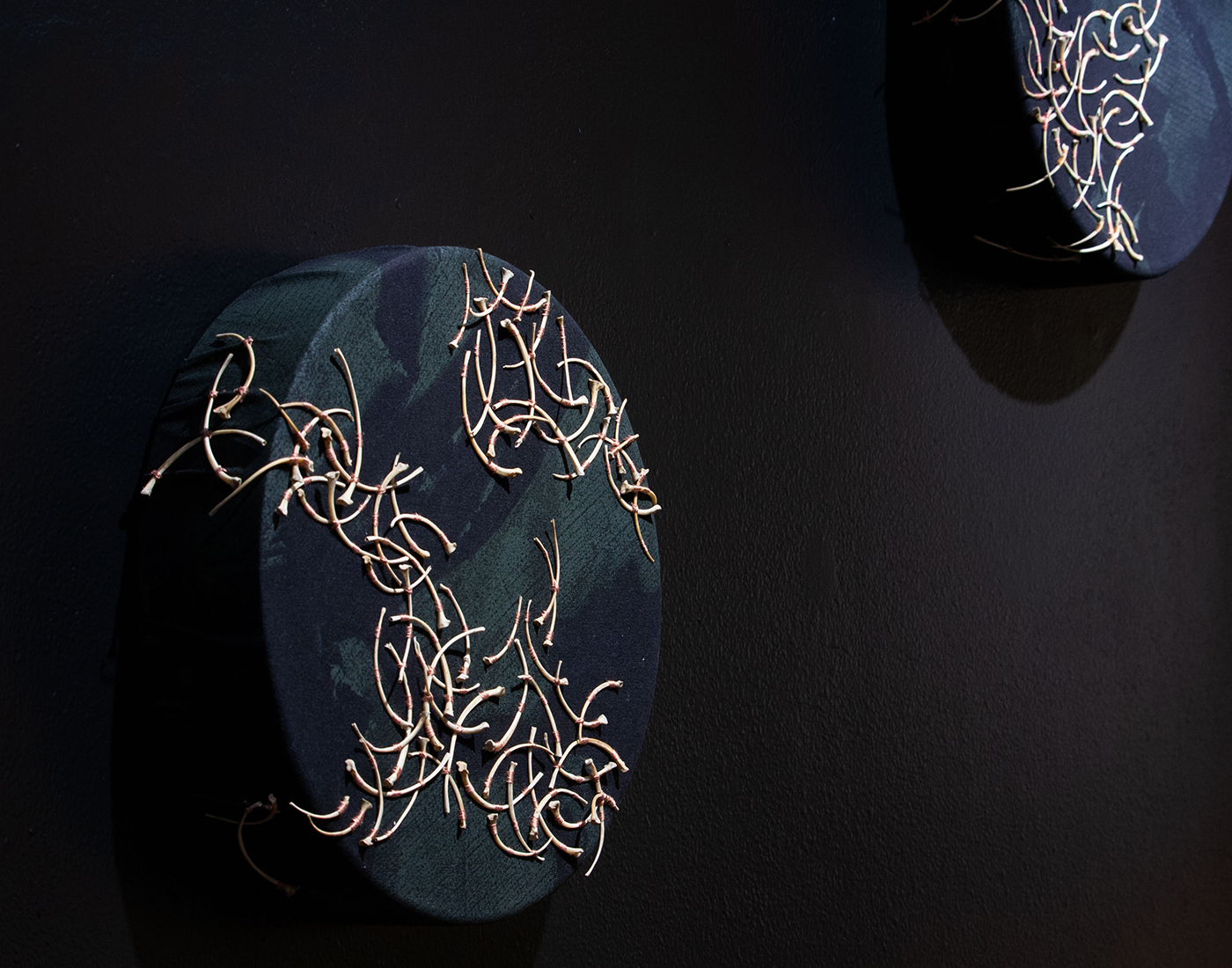 Two circular forms with hand-printed black fabric stretched over forms, embroidered with python bones. On dark background.