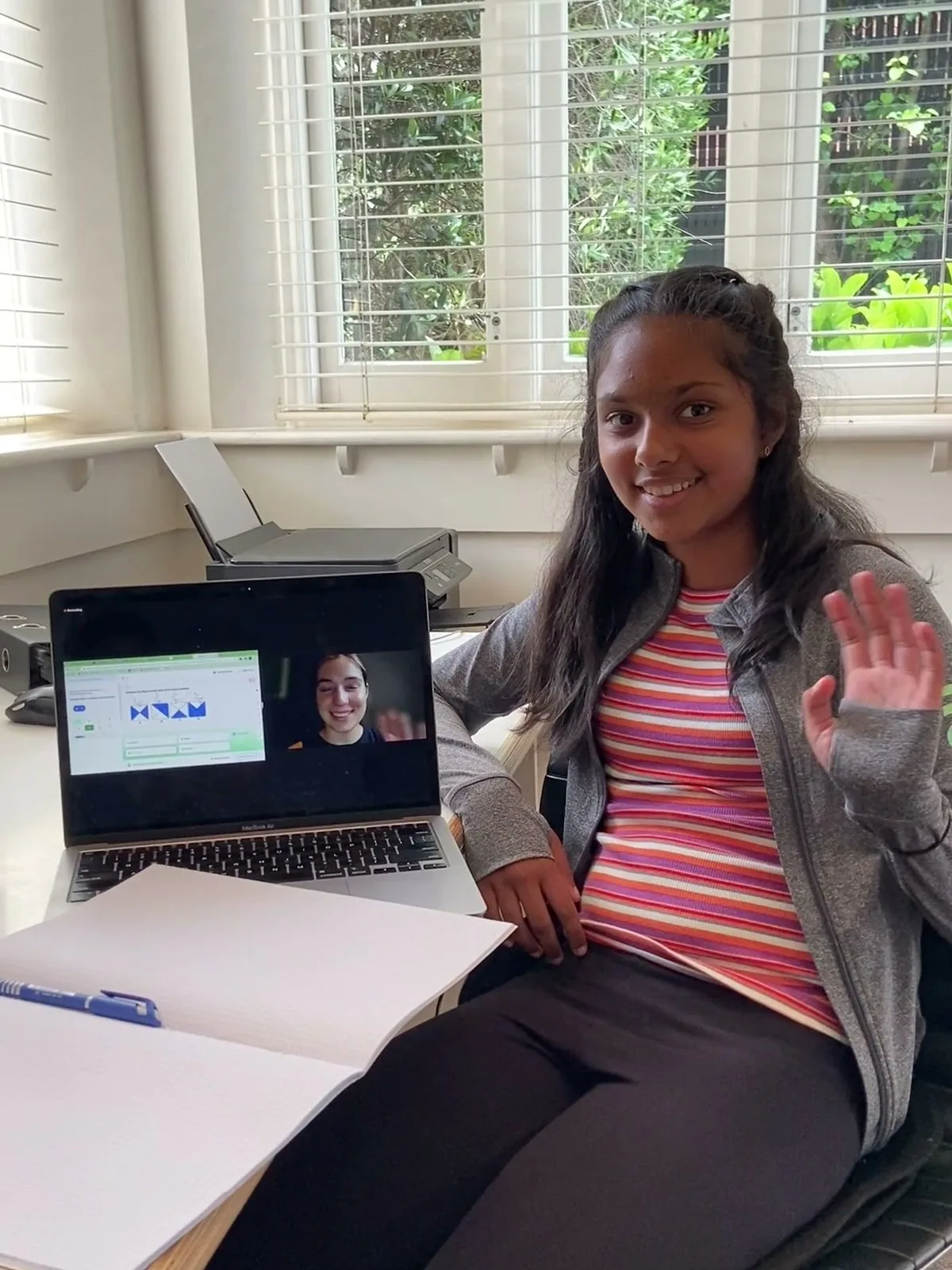 Student waving to her tutor at the start of an online lesson, the tutor live on her laptop screen