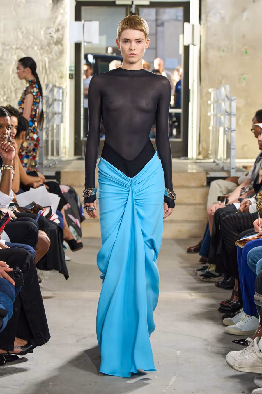 model walking down the runway wearing a sheer black turtleneck and flowy teal skirt with ruching.