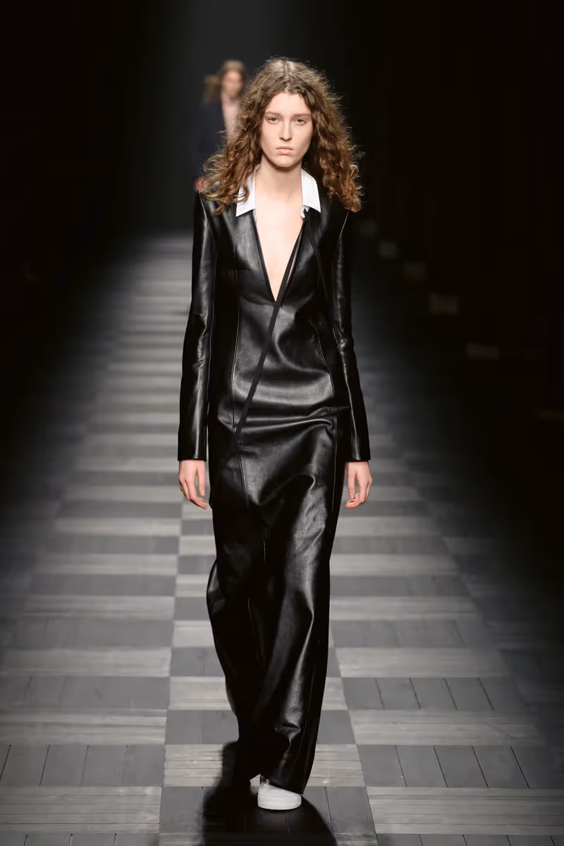 Ann Demeulemeester model walking the runway in straight leg black leather maxi dress with exposed cleavage.