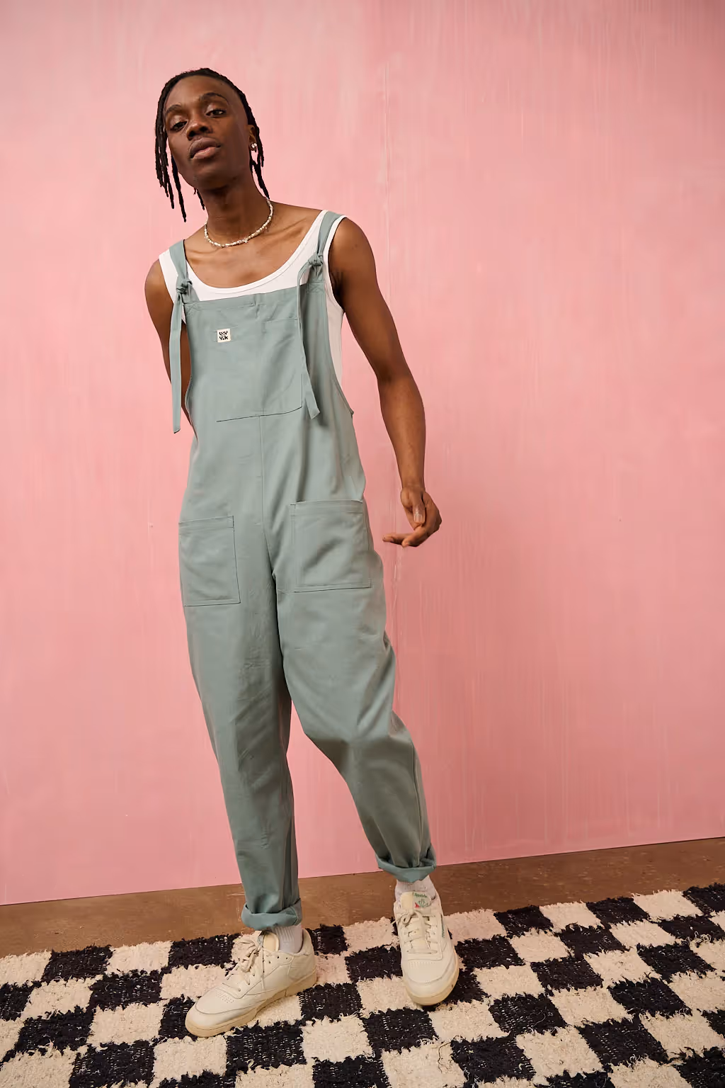 Male model wearing a Original Dungaree jumpsuit in Brighton blue over a tank top. He stands in front of a pink background.