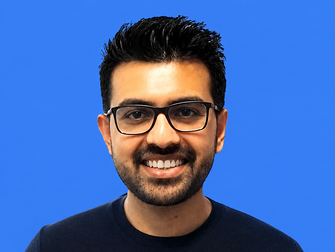Smiling man with black hair, glasses, and beard wearing a dark shirt against blue background.
