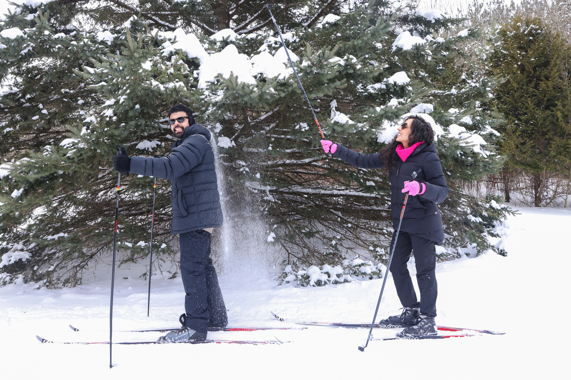 two people are cross-country skiing through the forest on a winter day. a girl is using her ski pole to hit the snow off the tree branches above to playfully dump snow on her friend.