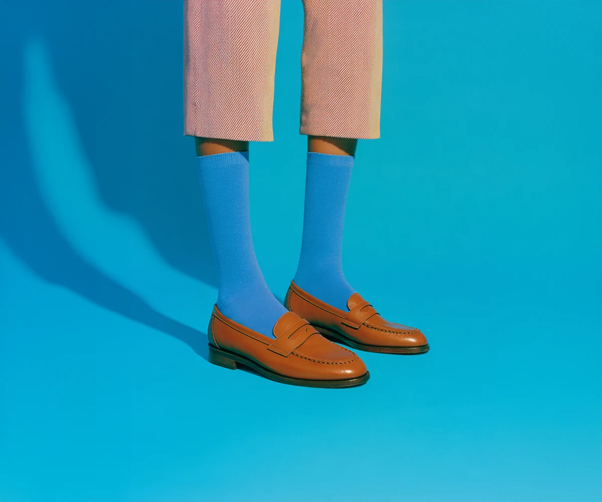 Person wearing beige cropped pants, bright blue socks, and brown leather loafers against a blue background.
