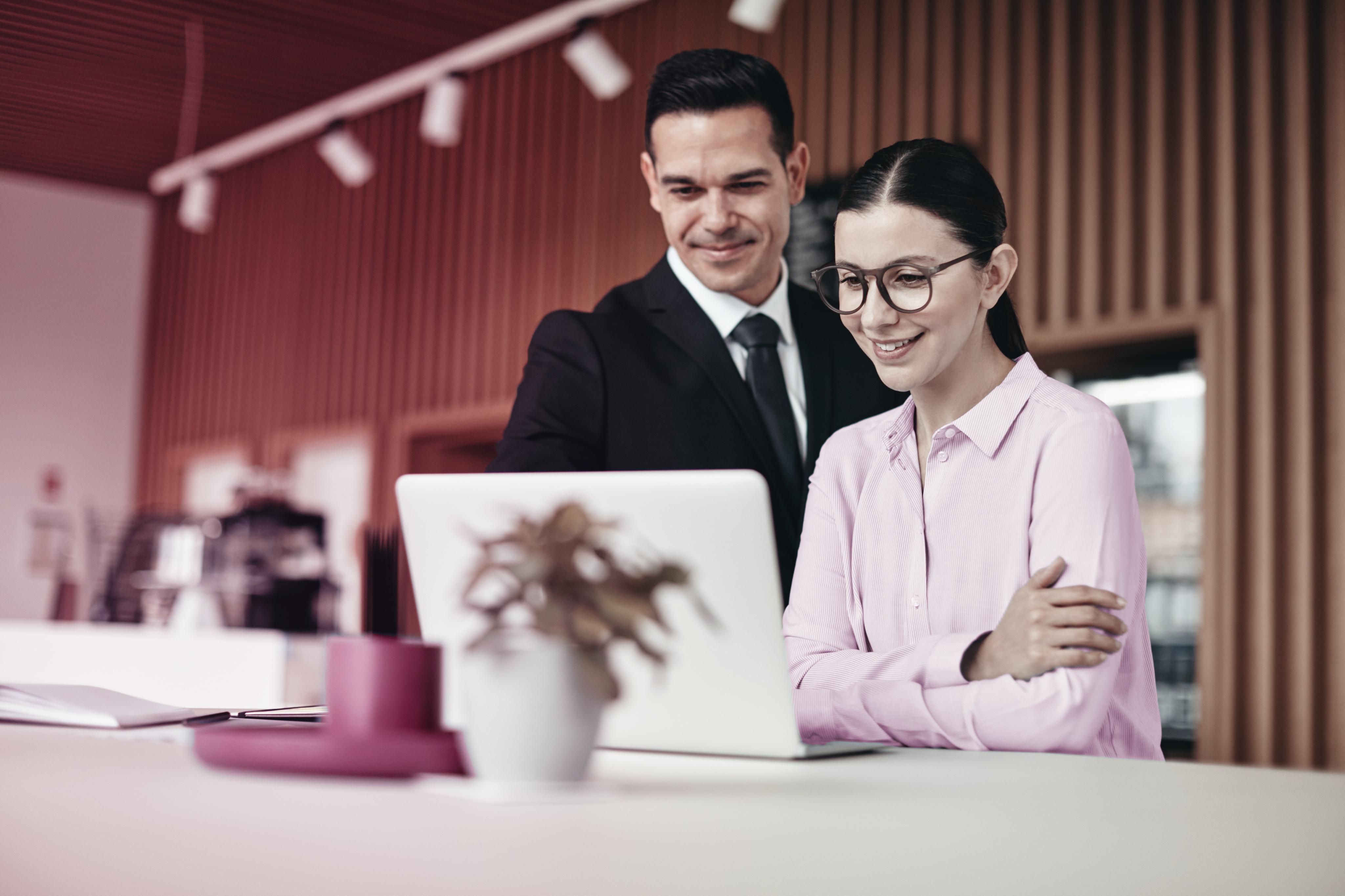 A imagem mostra uma mulher e um homem em um escritório olhando para um laptop. O homem está sorrindo e parece estar explicando algo à mulher. A mulher também parece contente e está com os braços cruzados. Eles estão juntos em um ambiente de trabalho, possivelmente discutindo um projeto ou uma tarefa.