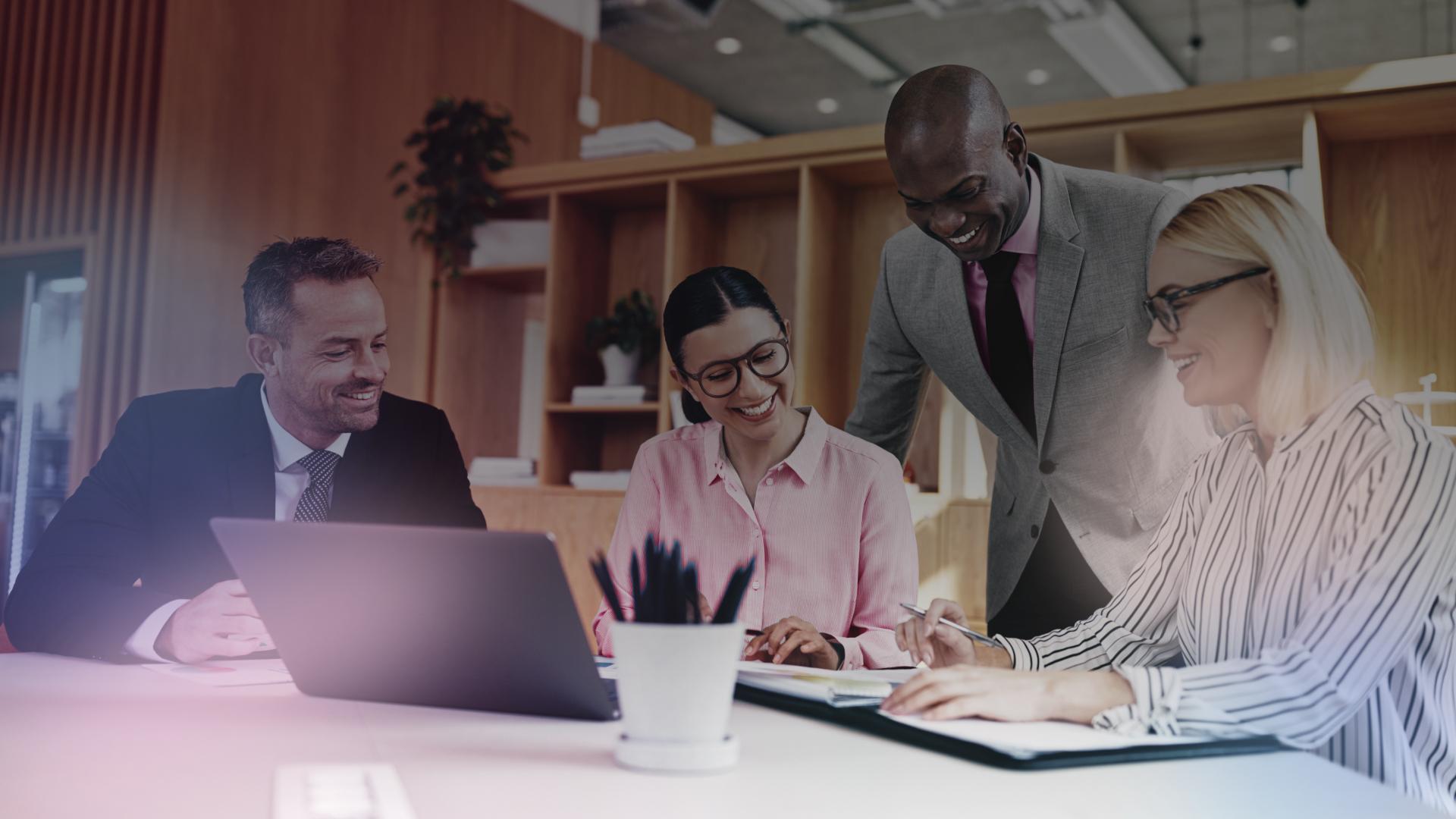 A imagem mostra um grupo de quatro colegas de trabalho em um escritório moderno. Eles estão reunidos em torno de uma mesa, olhando para um laptop. Há uma atmosfera positiva e de colaboração, com todos sorrindo e parecendo envolvidos na conversa.  A imagem sugere uma atmosfera de trabalho dinâmica e profissional.