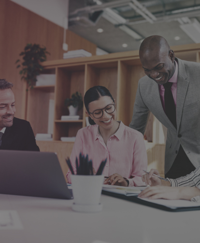 A imagem mostra um grupo de quatro colegas de trabalho em um escritório moderno. Eles estão reunidos em torno de uma mesa, olhando para um laptop. Há uma atmosfera positiva e de colaboração, com todos sorrindo e parecendo envolvidos na conversa.  A imagem sugere uma atmosfera de trabalho dinâmica e profissional.