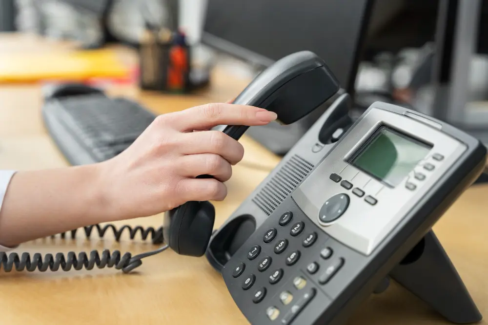 Person's hand lifting the receiver of a desk phone with a computer keyboard in the background.