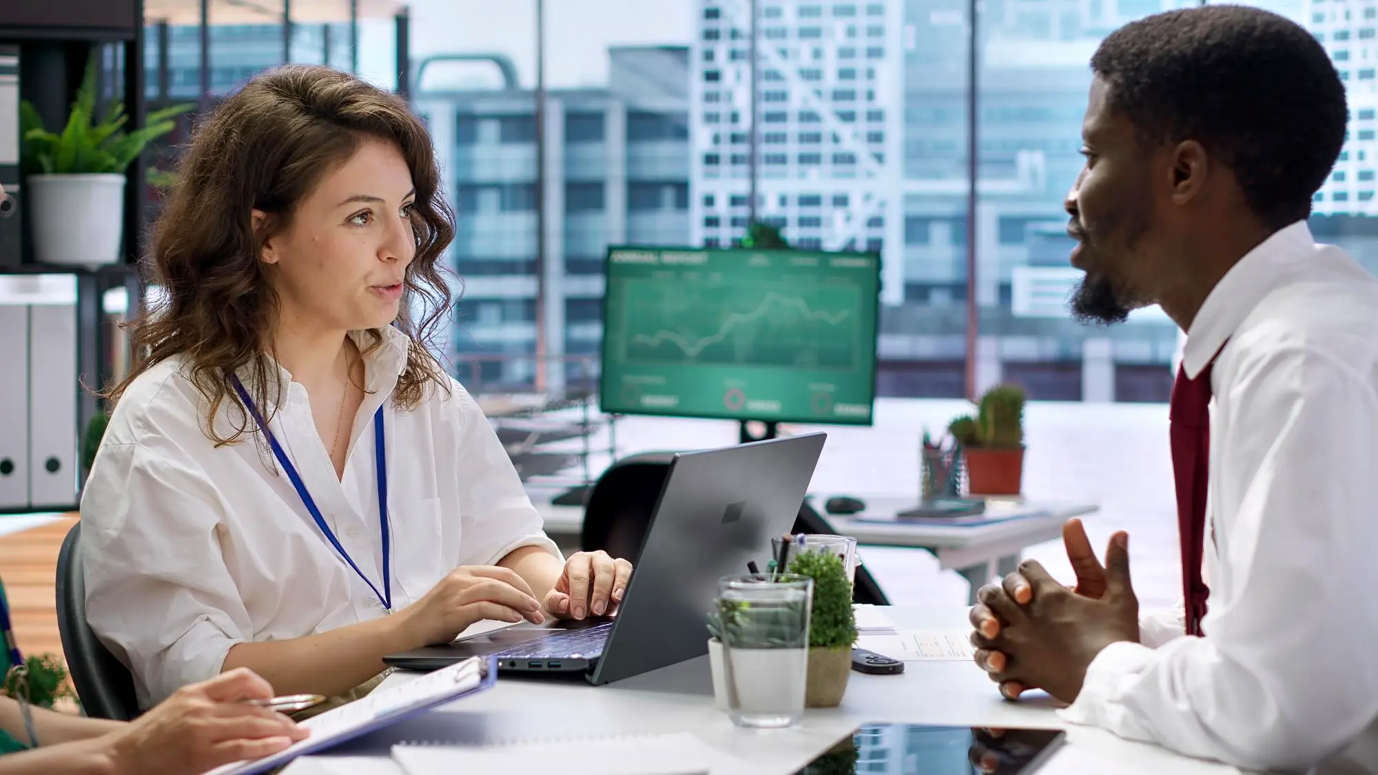 Man in a gray suit using a smartphone and working on a laptop with financial charts on the screen at a wooden table.
