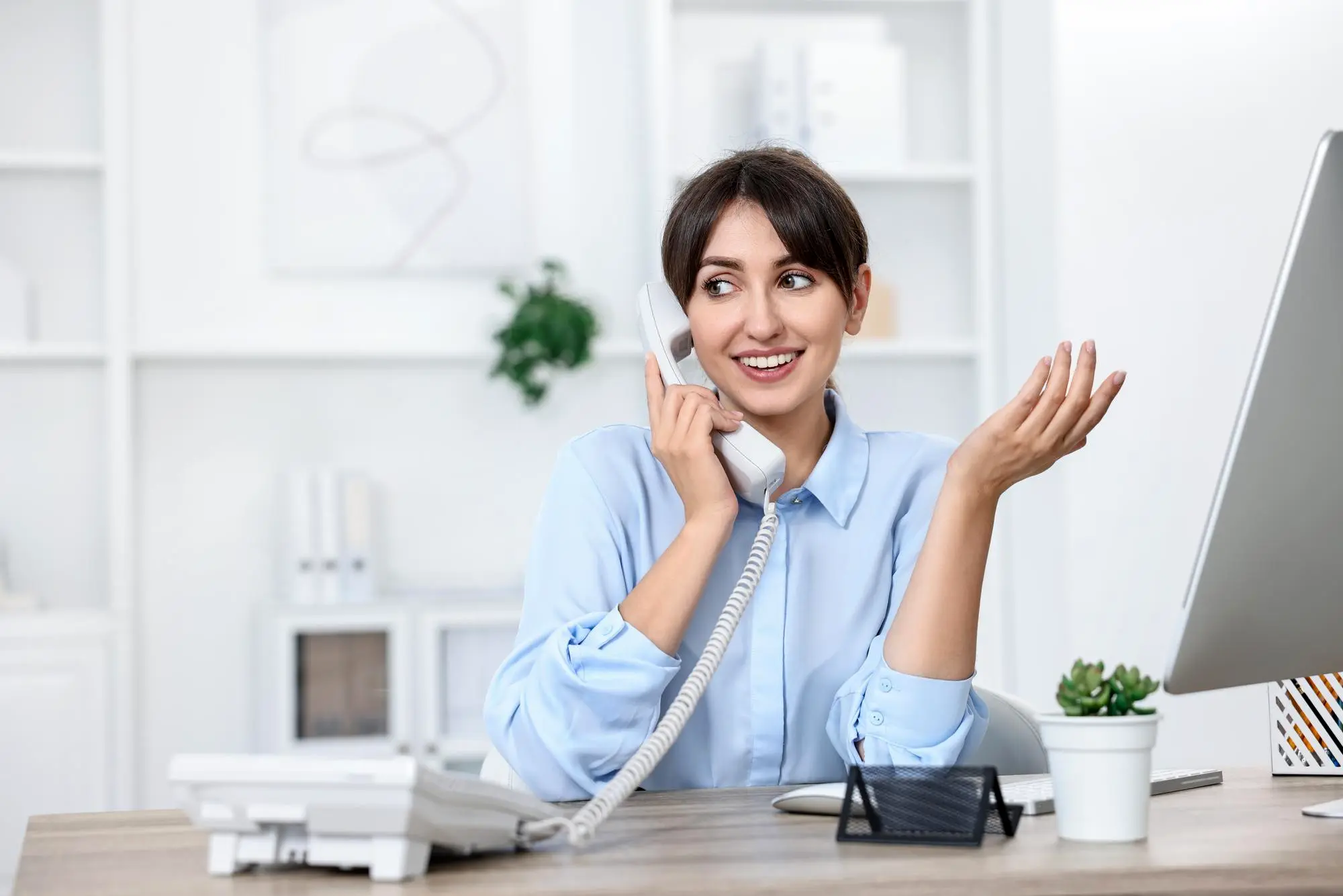 Smiling businesswoman in black suit talking on office desk phone while holding a pen.