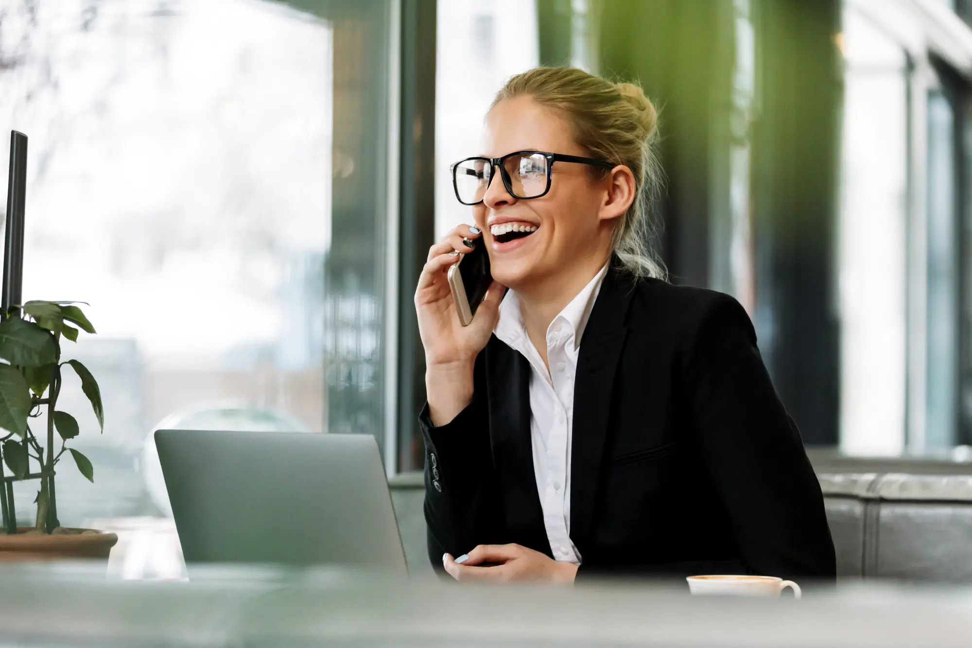 Man in a gray suit using a smartphone and working on a laptop with financial charts on the screen at a wooden table.