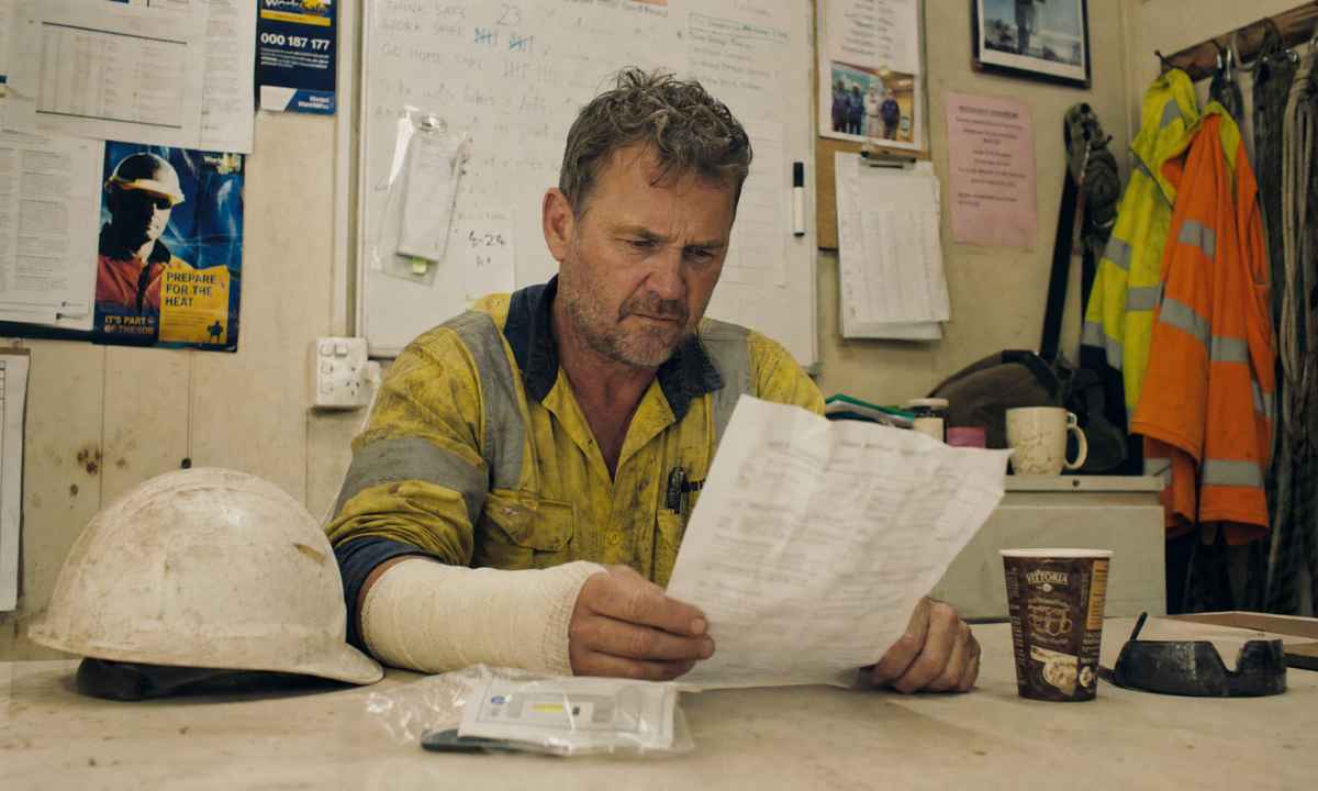Injured Queensland construction worker with bandaged arm reading WorkCover paperwork in a site office