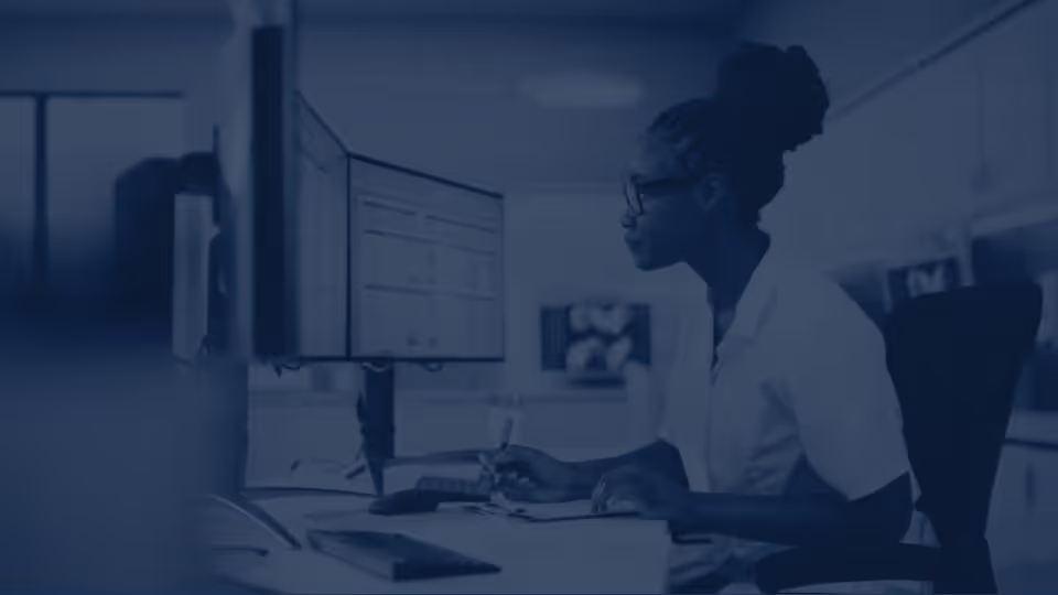 Professional woman wearing glasses, working at a desk with dual monitors and taking notes, in a modern office environment.