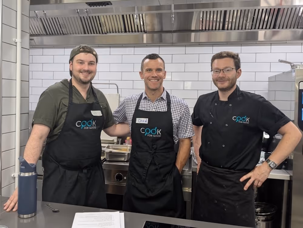 Three men in “Cook for Good” aprons stand behind a kitchen counter, posing for a group photo in a tiled commercial kitchen with visible cooking equipment and utensils.
