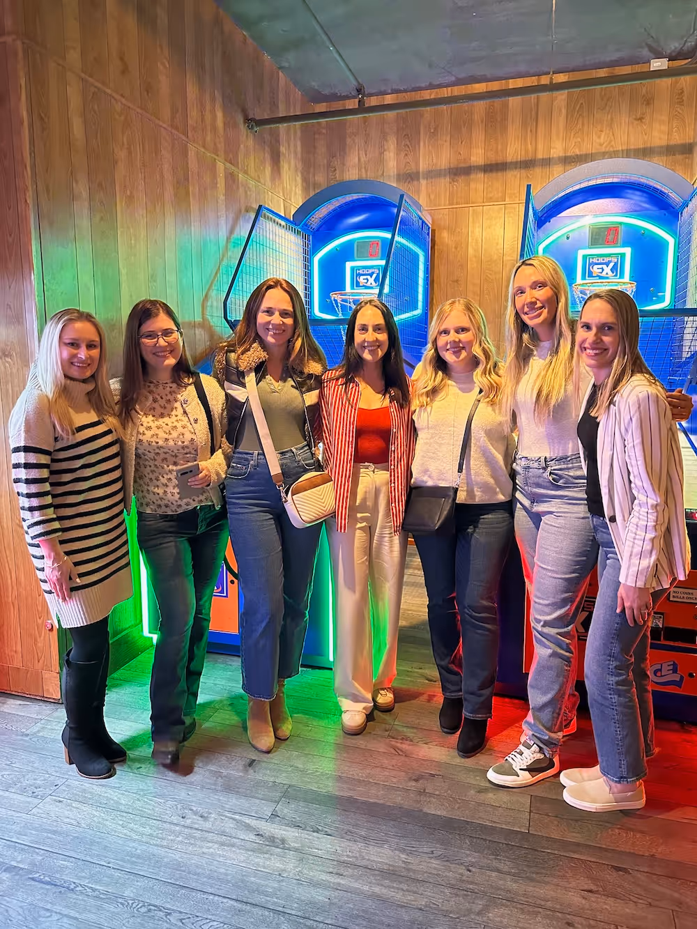 A group of seven women posing and smiling in front of brightly lit arcade basketball games during a casual team social or outing.