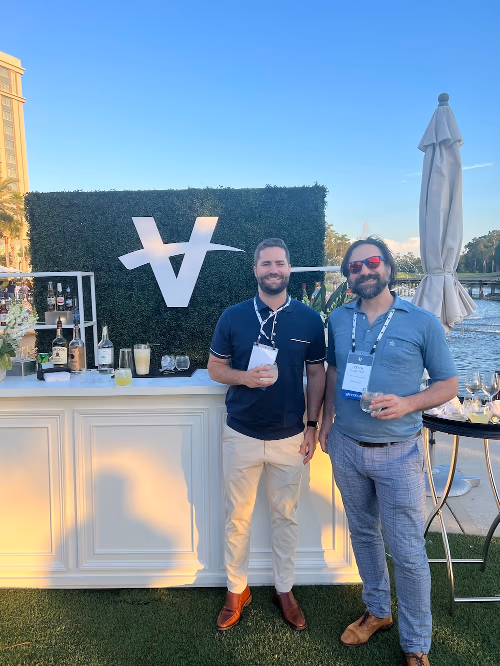 Two men pose with drinks in front of a branded outdoor bar setup at a sunny networking event, with a hedge wall featuring a large stylized "V" logo and bottles arranged on the counter.