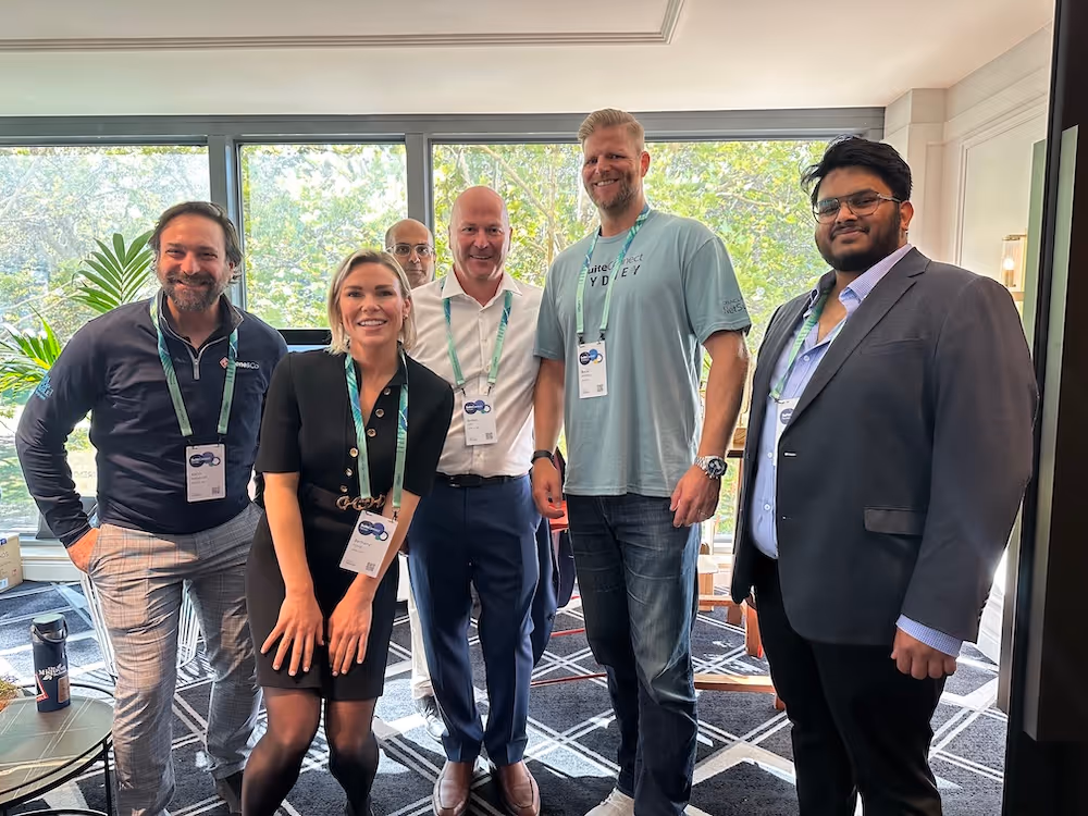 Six professionals wearing conference lanyards gather for a casual group photo in a well-lit venue, standing in front of windows with greenery outside.