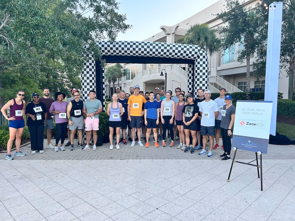 Large group of participants posing at the starting line of a 5K fun run and walk event, with a checkered inflatable arch and a Zone & Co-branded sign in the foreground.