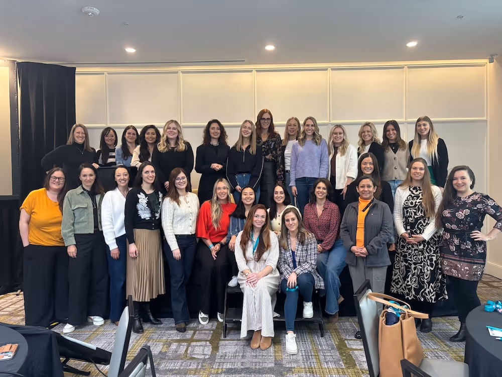 A large group of professionally dressed women pose together for a group photo in a conference or event space, many of them smiling in multiple rows.