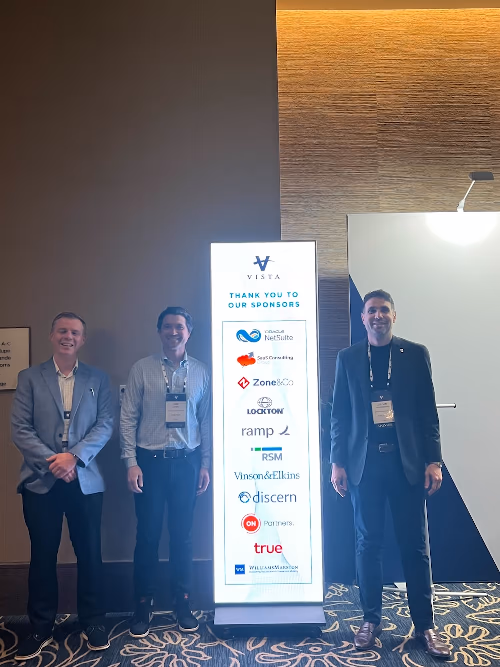 Three men standing next to a vertical digital sign displaying sponsor logos at a conference, including Zone & Co and other companies like NetSuite and RSM.