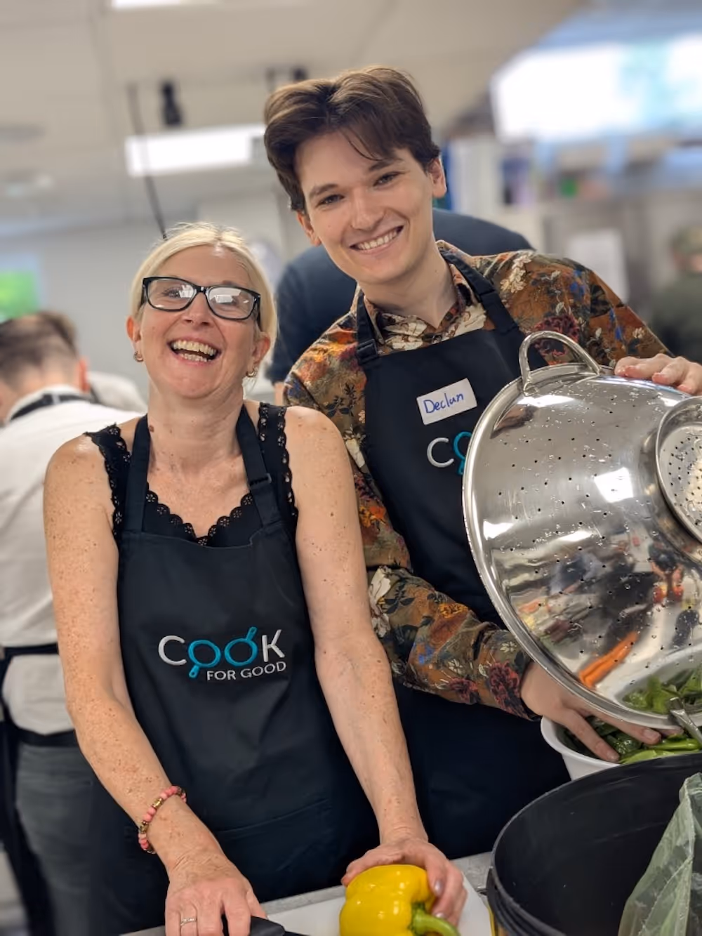 Two volunteers, one holding a yellow bell pepper and the other a large colander of vegetables, smile widely while participating in a Cook for Good charity cooking session.