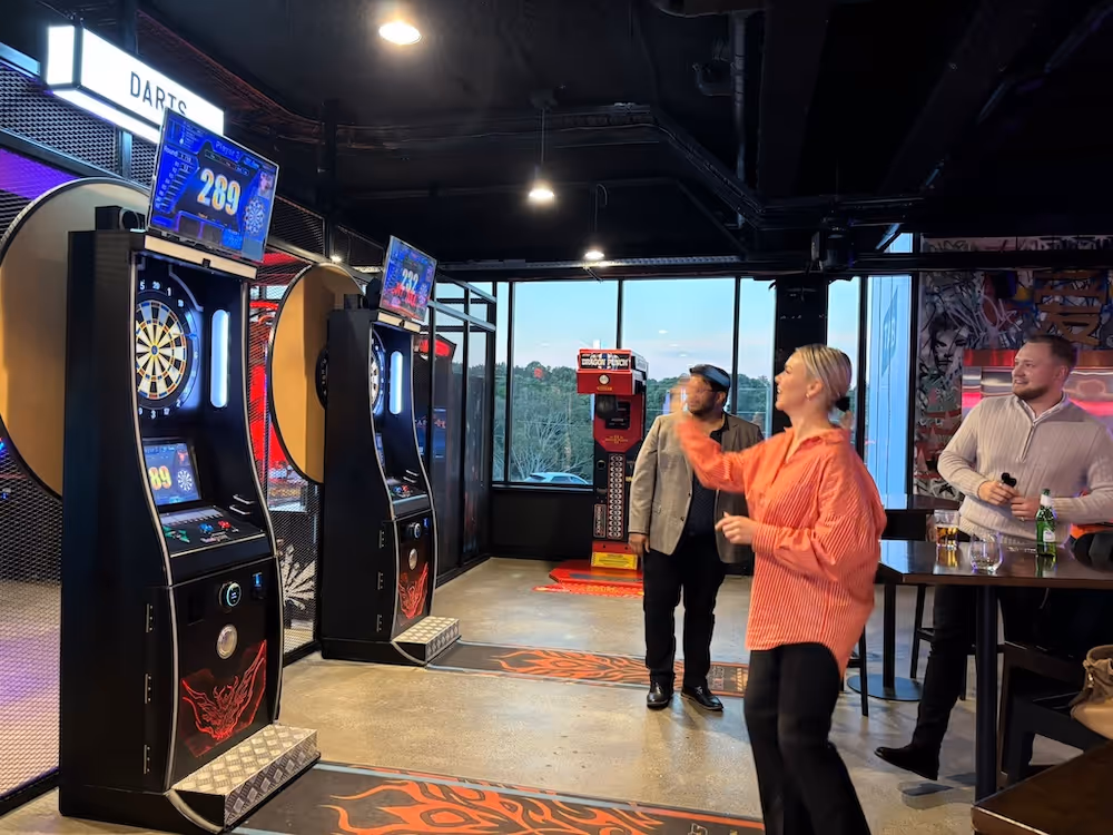 People enjoying a darts game in a modern arcade-style venue, with one woman mid-throw and two men watching in the background, surrounded by dart machines and drinks on a high table.