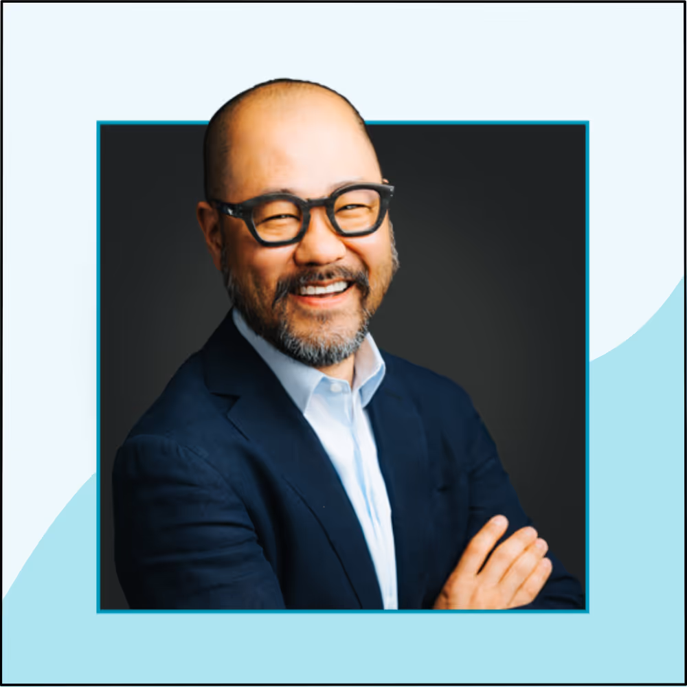 Smiling bald man with glasses and beard wearing a dark suit and light blue shirt against a dark background.