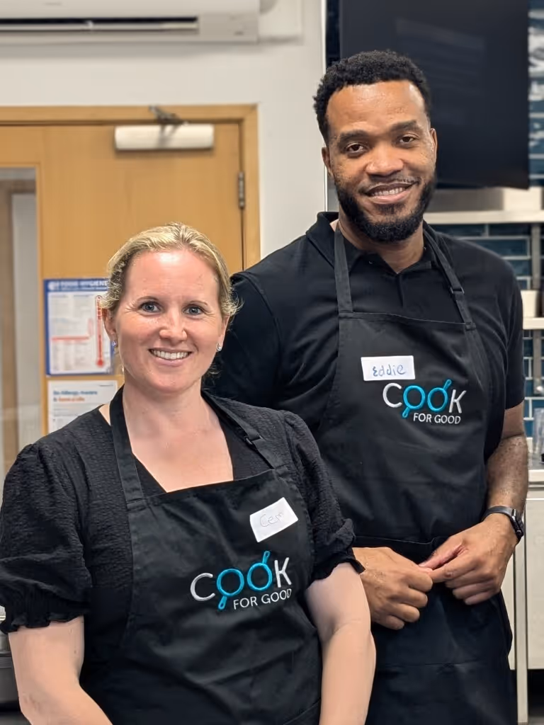 Two smiling people wearing black aprons with 'Cook for Good' logo and name tags in a kitchen setting.