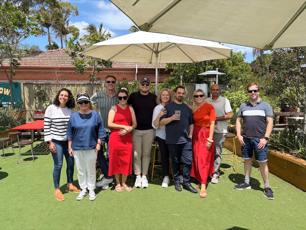 Group of ten people standing outdoors on green artificial turf under large umbrellas on a sunny day.