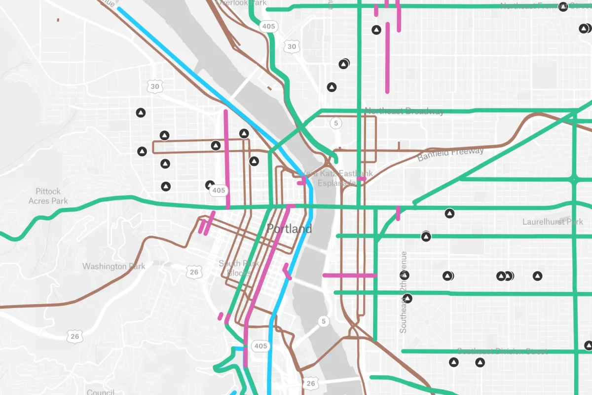 A simplified transit and road network map of Portland, Oregon, showing color-coded routes and infrastructure markers across the city's grid and Willamette River crossings to assist in urban mobility planning.