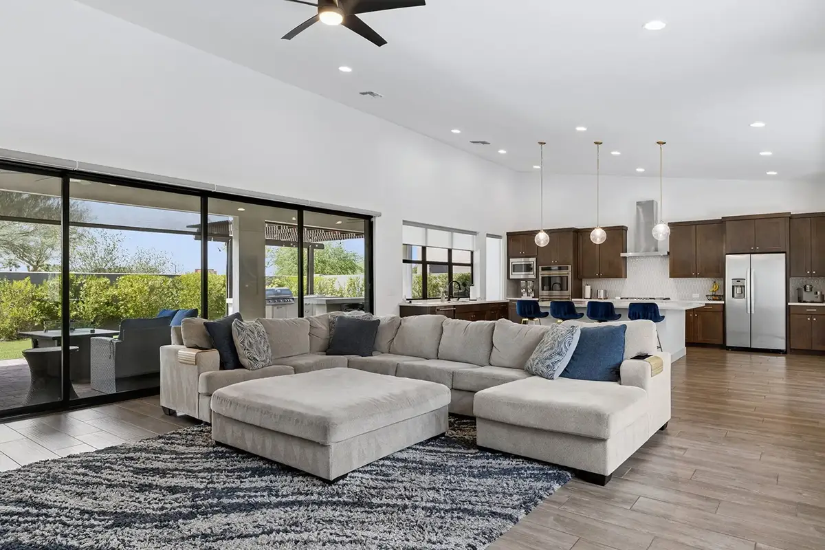 Open-plan living area with large sectional sofa, vaulted ceiling with ceiling fan, floor-to-ceiling sliding glass doors opening to a backyard patio, and a kitchen with dark wood cabinetry, island with blue bar stools, and stainless steel appliances
