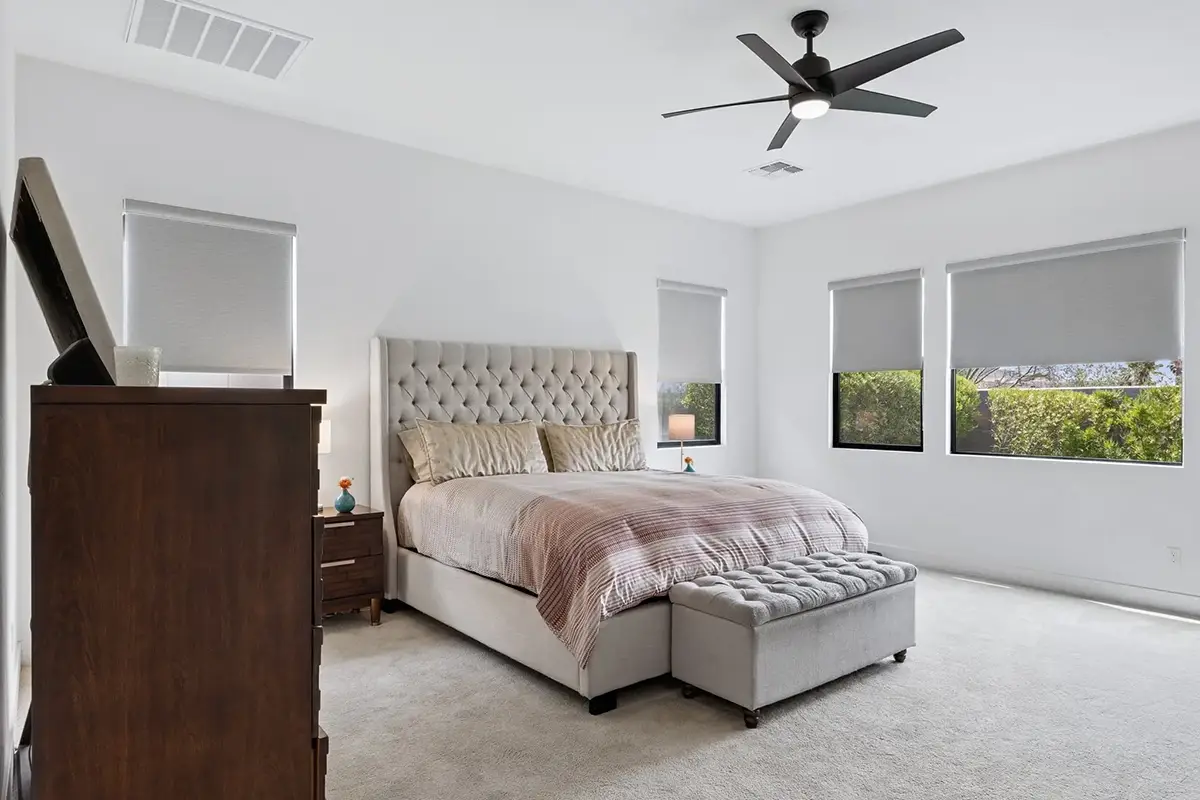 Primary bedroom with tufted upholstered bed, storage bench, dark wood dresser, ceiling fan, and multiple windows with roller shades