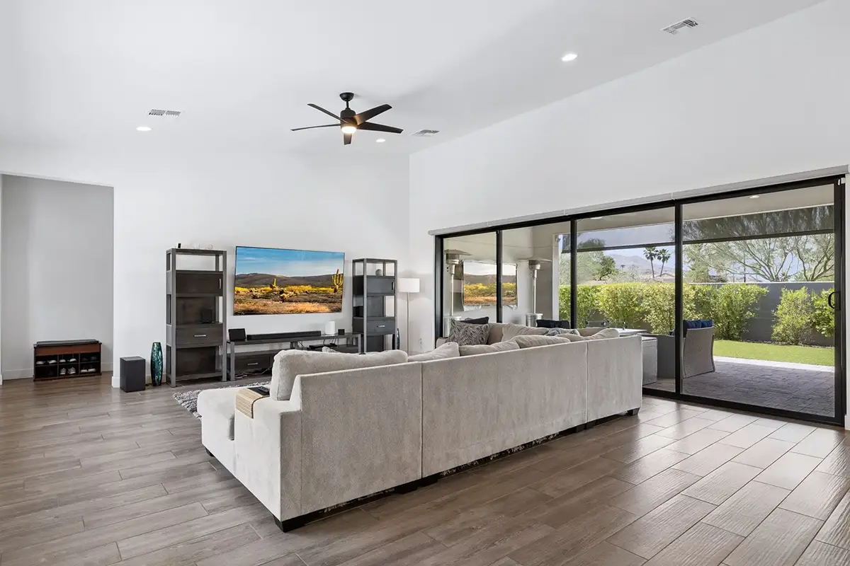 Open living room with sectional sofa, large TV, media shelving, ceiling fan, and sliding glass doors opening to a green backyard