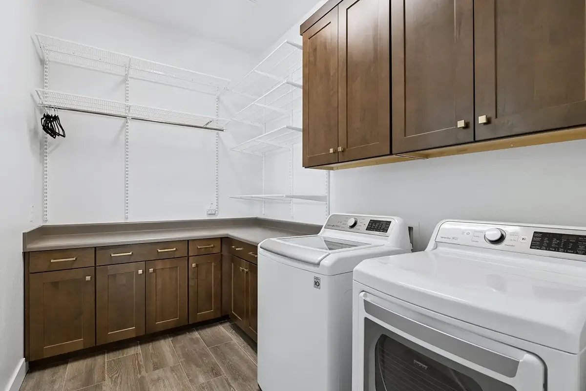 Spacious laundry room with top-load washer and dryer, dark wood cabinetry with gold hardware, L-shaped laminate countertop, upper wall cabinets, white wire shelving system with hanging rod, and wood-look tile flooring