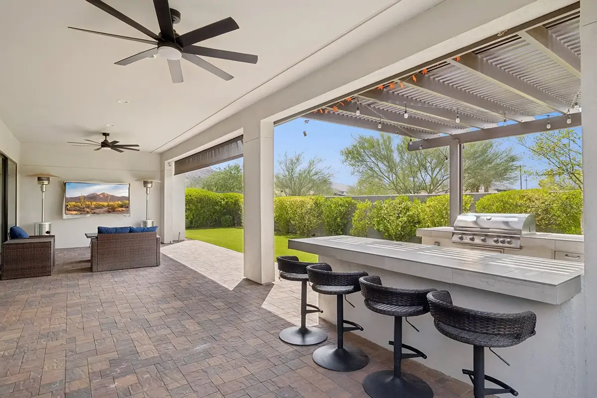 Covered backyard patio with built-in outdoor kitchen, white stone bar counter with three wicker bar stools, stainless built-in grill, louvered pergola with string lights, ceiling fans, outdoor TV, wicker lounge seating with blue cushions, and lush green he
