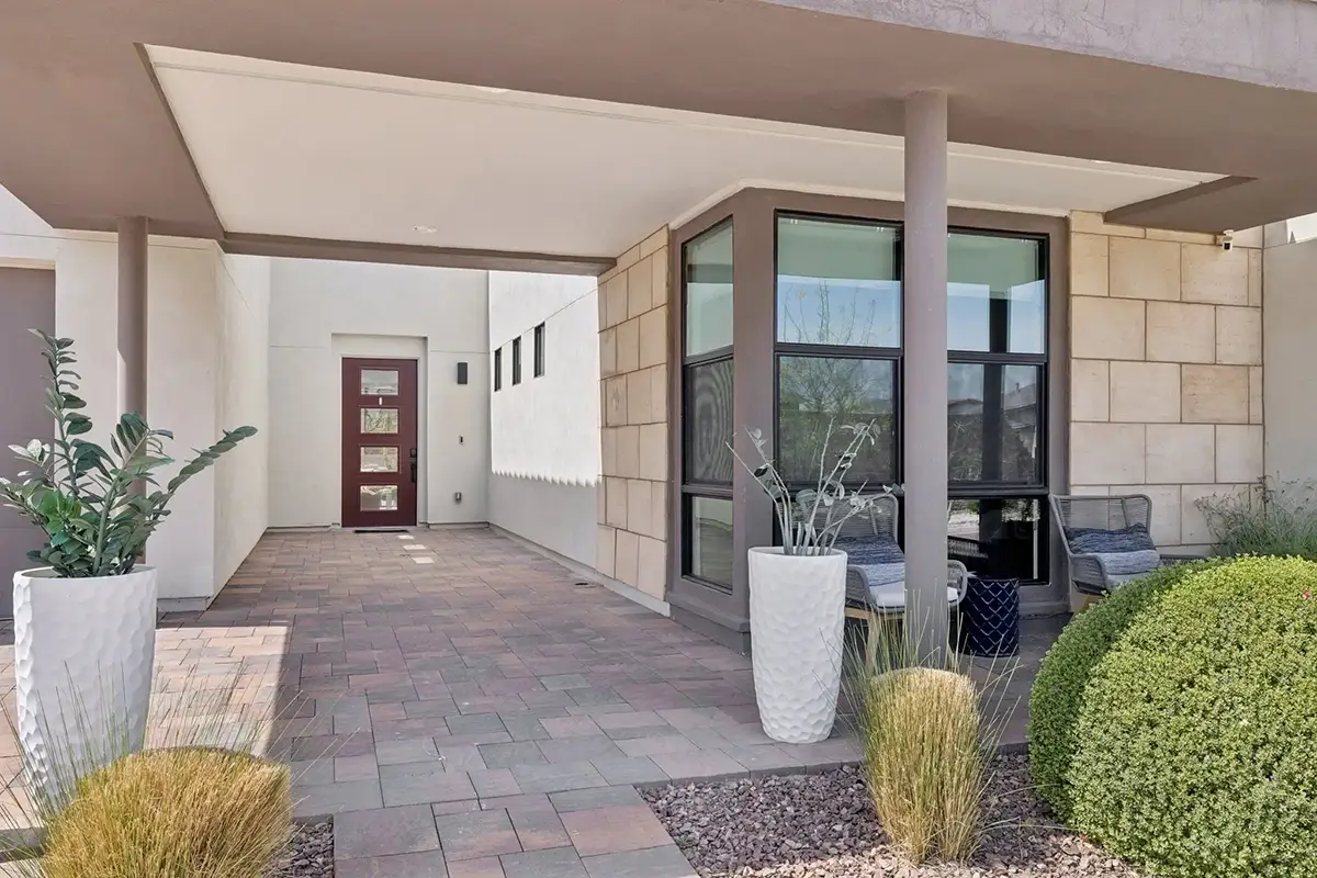 Covered front entry walkway with brick paver flooring, stone-clad exterior walls, dark-framed floor-to-ceiling windows, deep red front door with glass panels, tall white planters with greenery, and outdoor seating area with desert landscaping