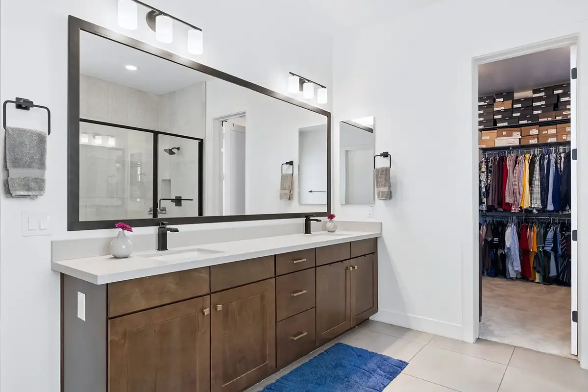 Primary bathroom with double vanity, dark wood cabinetry, white quartz countertop, matte black fixtures, large framed mirror with vanity lighting, blue bath mat, and open doorway to a walk-in closet with organized clothing and stacked shoeboxes