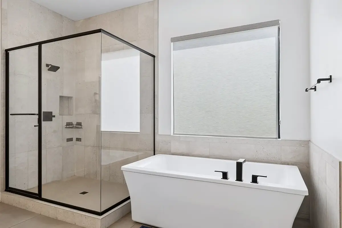 Primary ensuite bathroom with freestanding white soaking tub and matte black fixtures, large glass-enclosed walk-in shower with black frame, travertine tile walls and flooring, sheer roller shade window, and wall-mounted faucet fitting