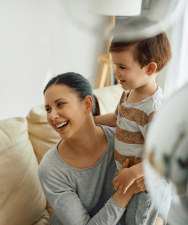Une femme aux cheveux longs tenant un enfant en bas âge tout en tenant une tasse blanche, avec un homme en arrière-plan lors d'une journée ensoleillée.