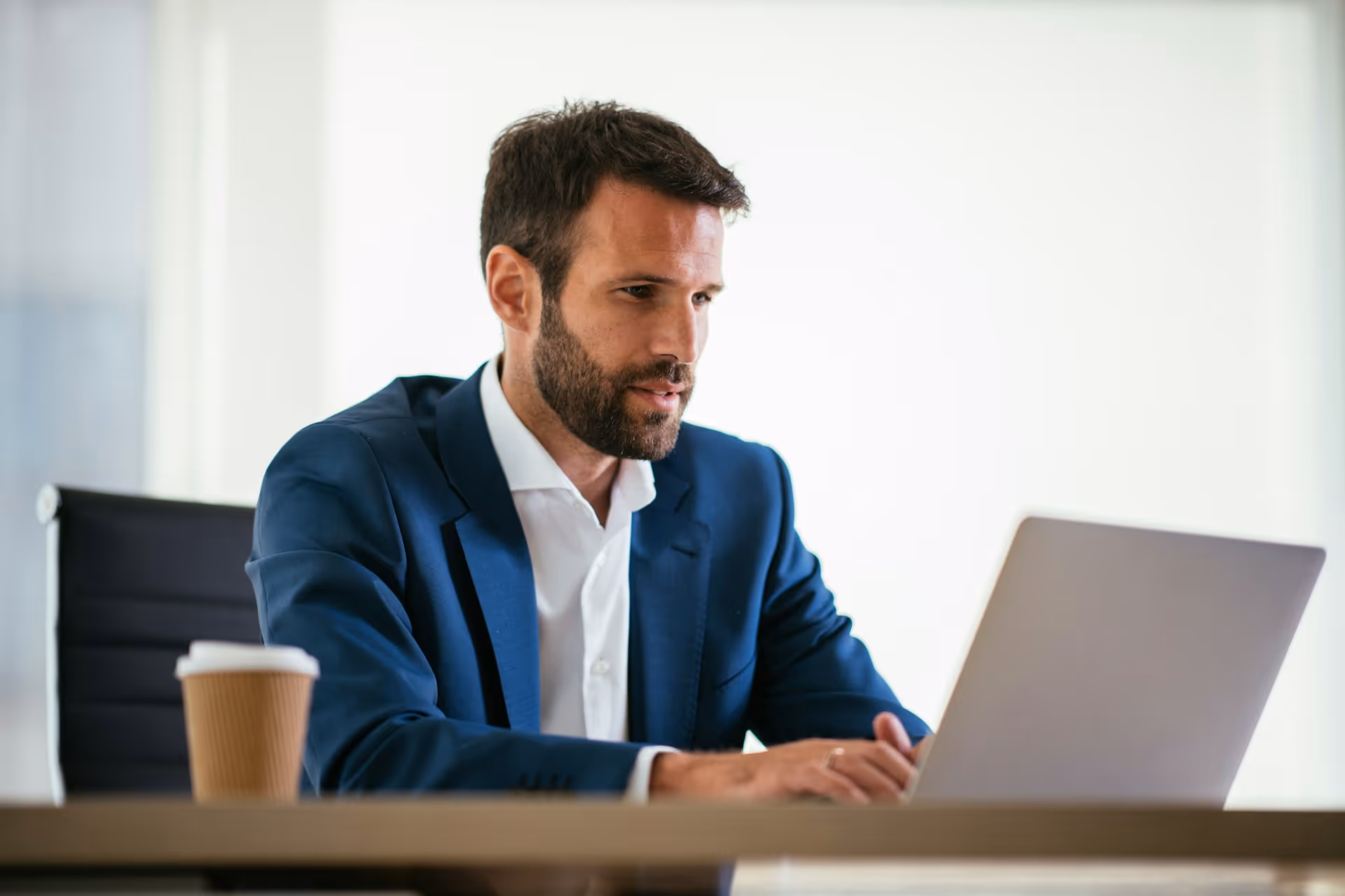 Homme d'affaires en costume bleu travaillant sur un ordinateur portable avec une tasse de café à côté.