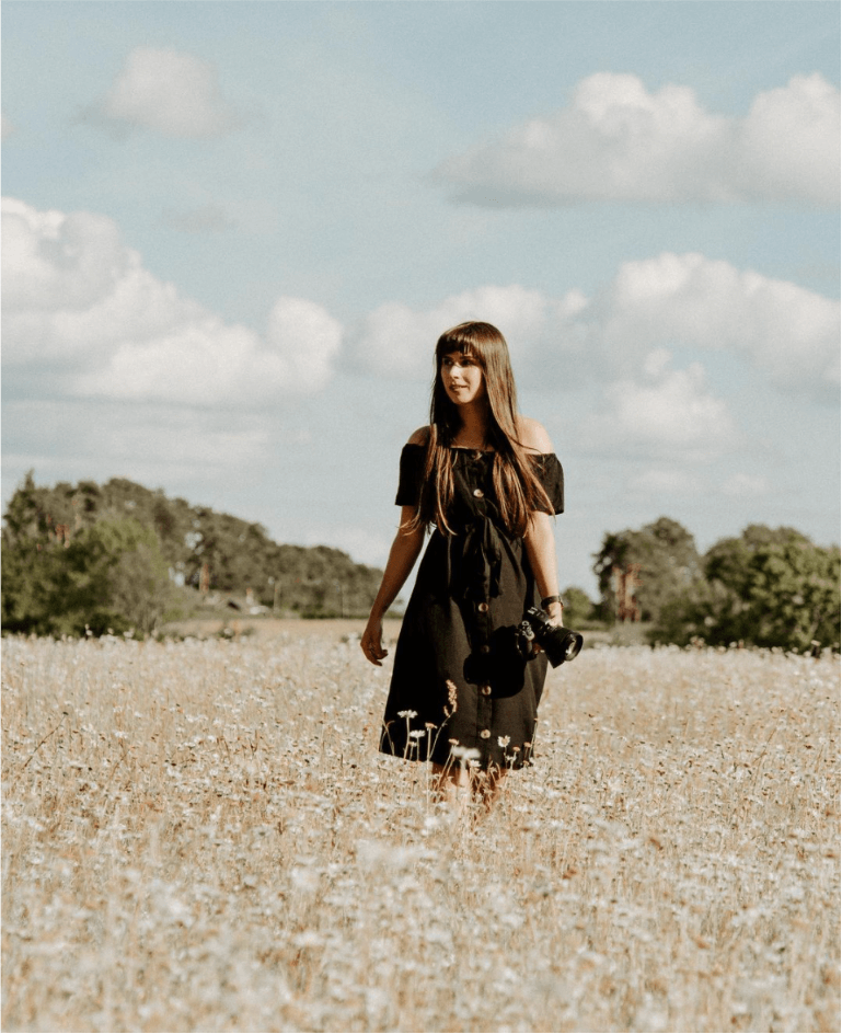 A photo of Buckinghamshire based wedding and studio photographer, Gemma Randall standing in the middle of a golden field with a camera.