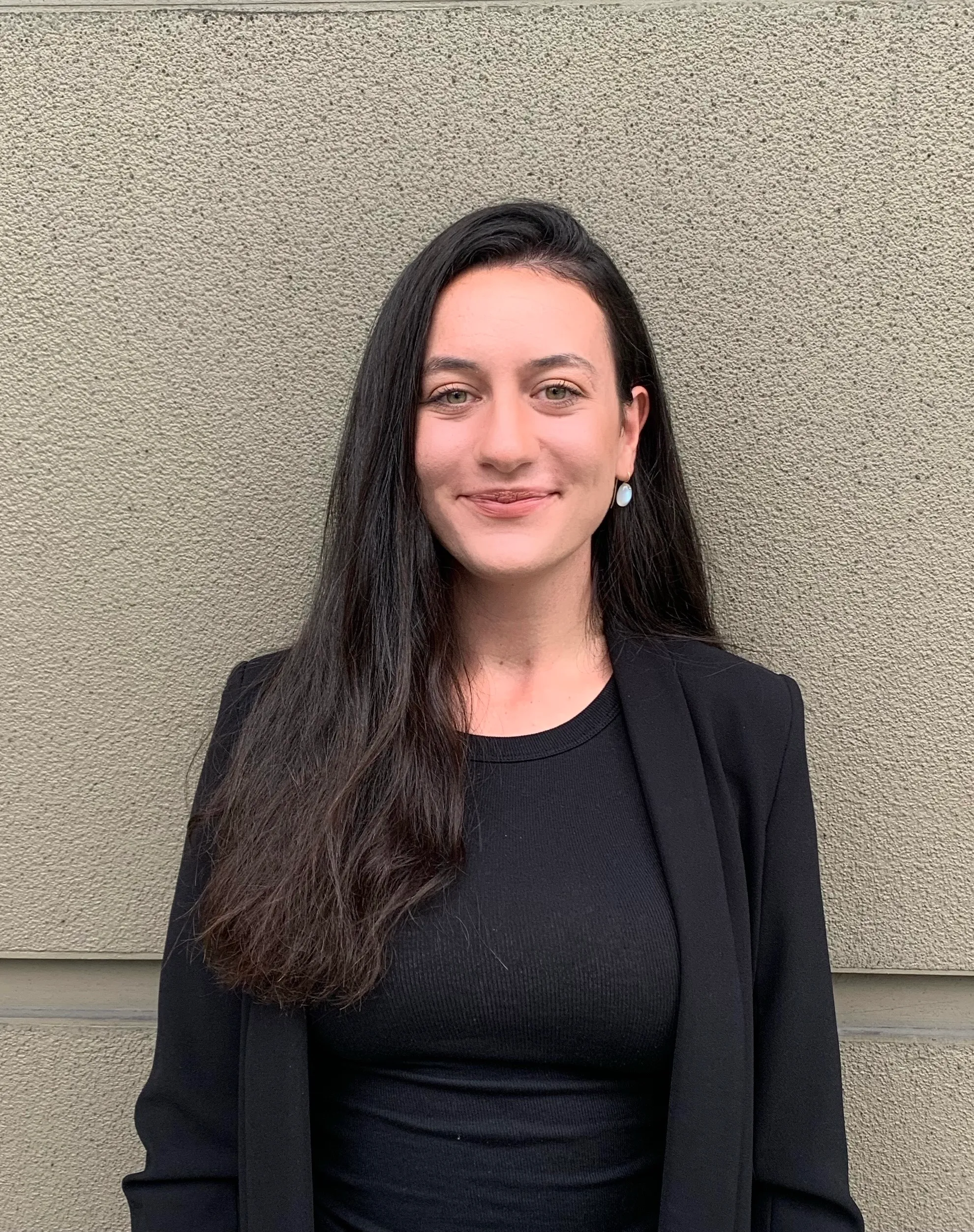Smiling woman with long dark hair wearing a black blazer and shirt standing against a textured beige wall.