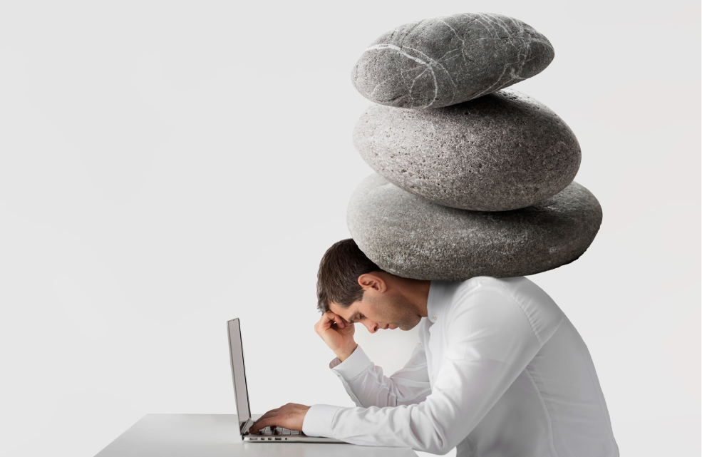 Stressed man at laptop with large stones stacked on his back, symbolizing overwhelming pressure and emotional burden
