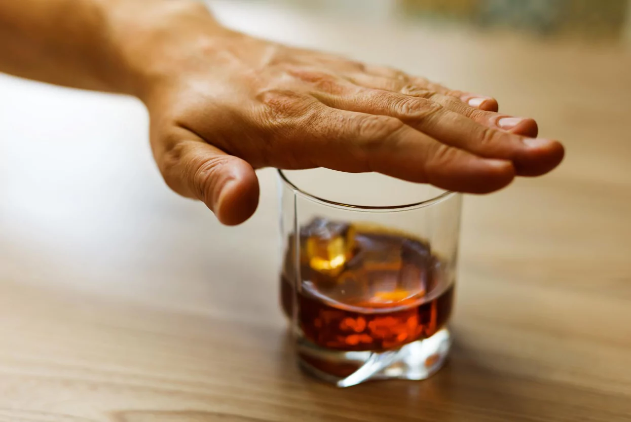 A person's hand reaching out to refuse a glass of whiskey with an ice cube on a wooden table.