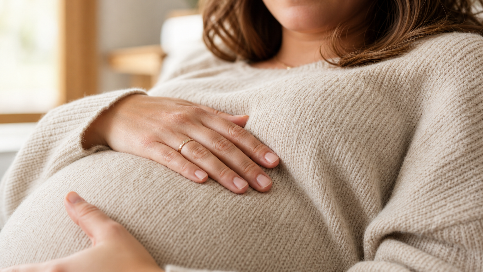A pregnant woman in her early 30s rests both hands on her belly over a knit sweater in warm window daylight, her calm face softly visible above.