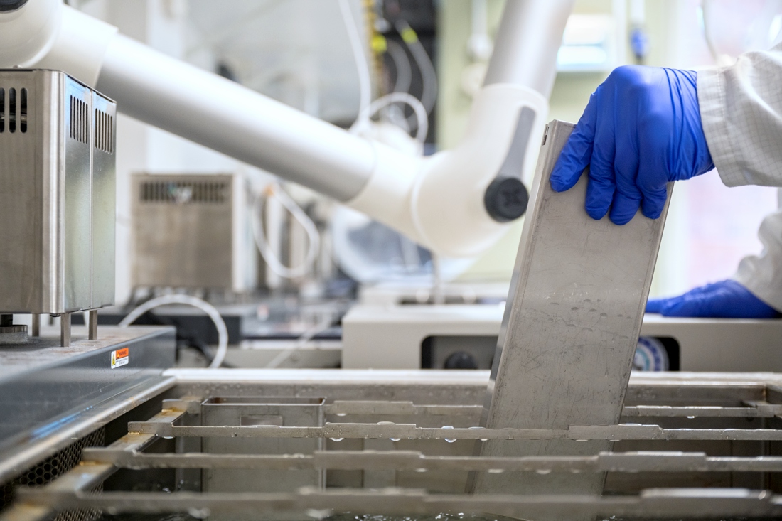 A person wearing blue gloves and a lab coat is handling a large metal plate, partially immersing it into an industrial liquid tank in a laboratory.