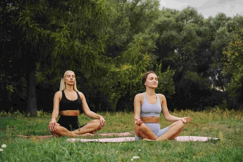 Two women breathe in seated yoga pose outside on grass