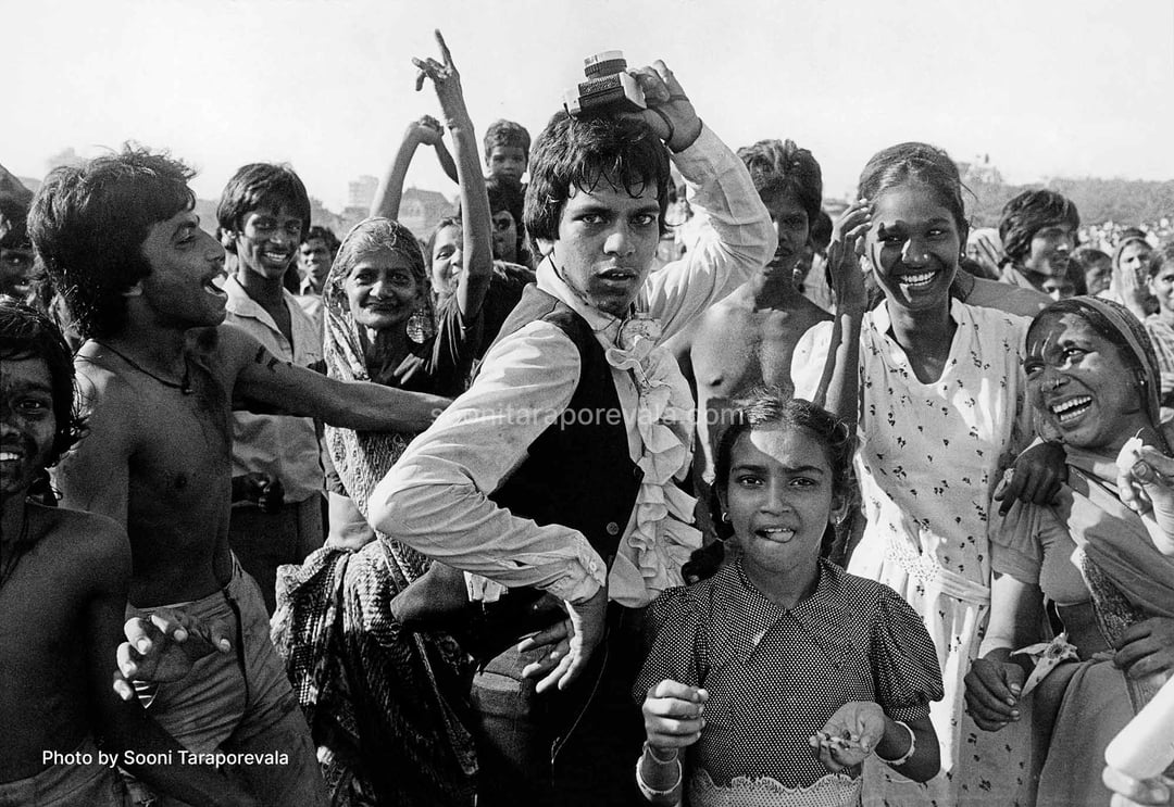 Dense black-and-white crowd at Holi, faces smeared with colour, a young man in a frilled shirt and waistcoat mid-dance holding a camera above his head, laughing women on either side