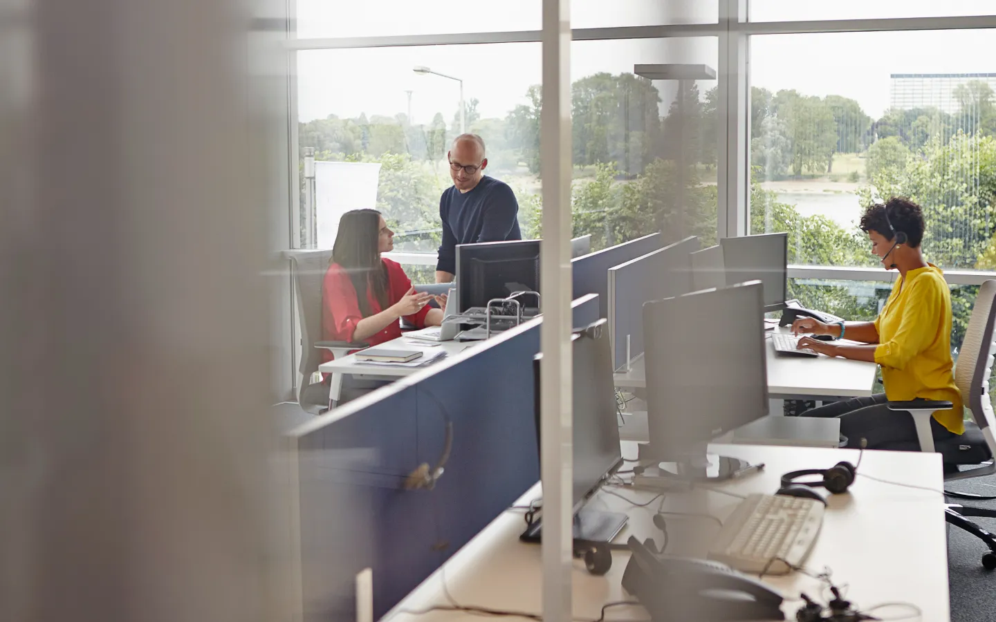 Office workspace with people working at desks, one woman in yellow using headset and two colleagues discussing near computer.